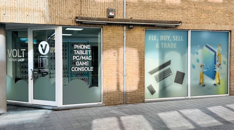 De glazen hoekgevel van Volt Repair Shop: het V-logo en de openingsuren op de deur, 'phone, tablet, PC/Mac, game, console' op het raam en de 'fix, buy, sell & trade'-etalage ernaast.